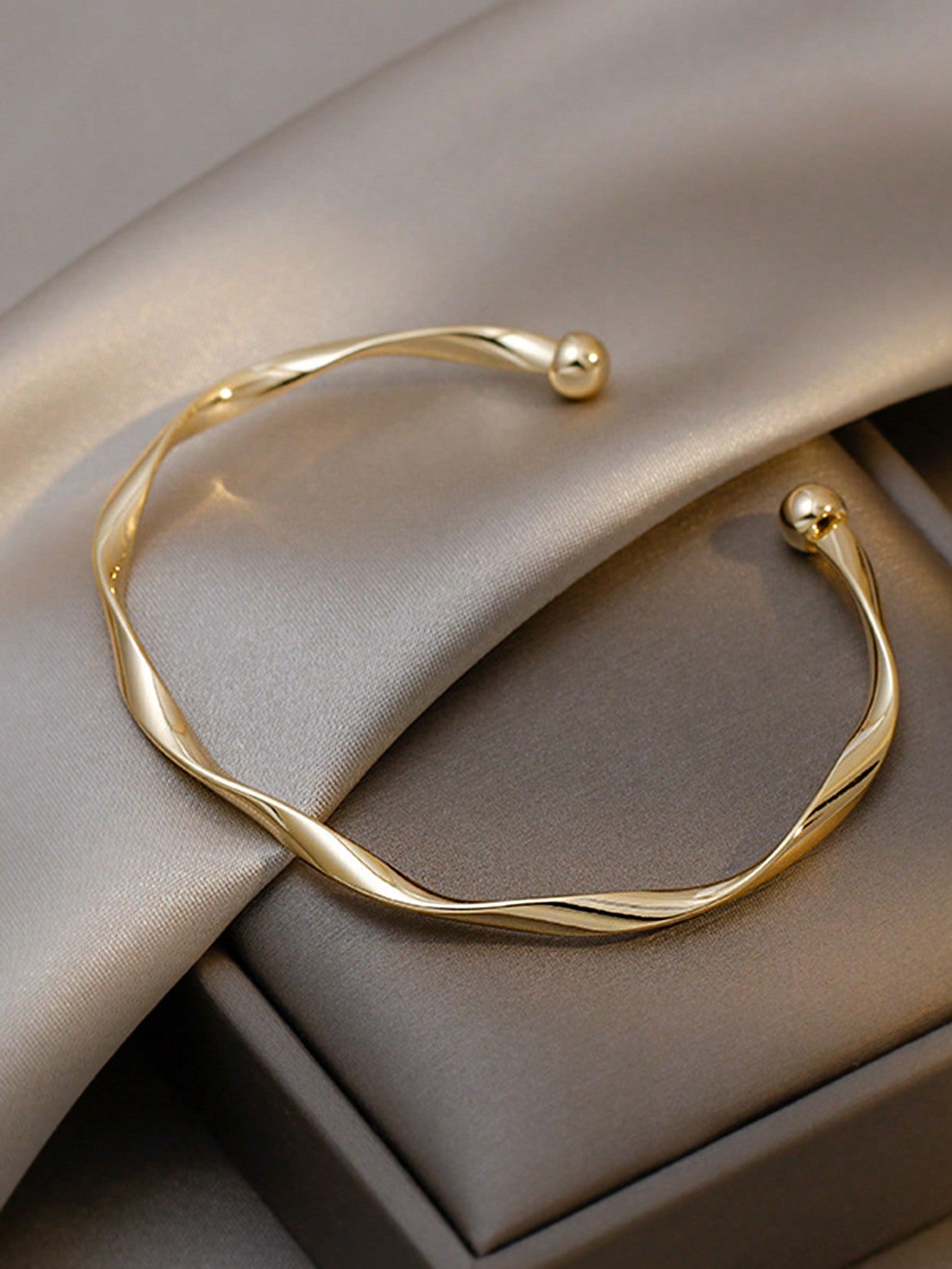Gold twisted bangle bracelet with a ball accent on one end. Elegant, minimalist design displayed on a gray fabric background