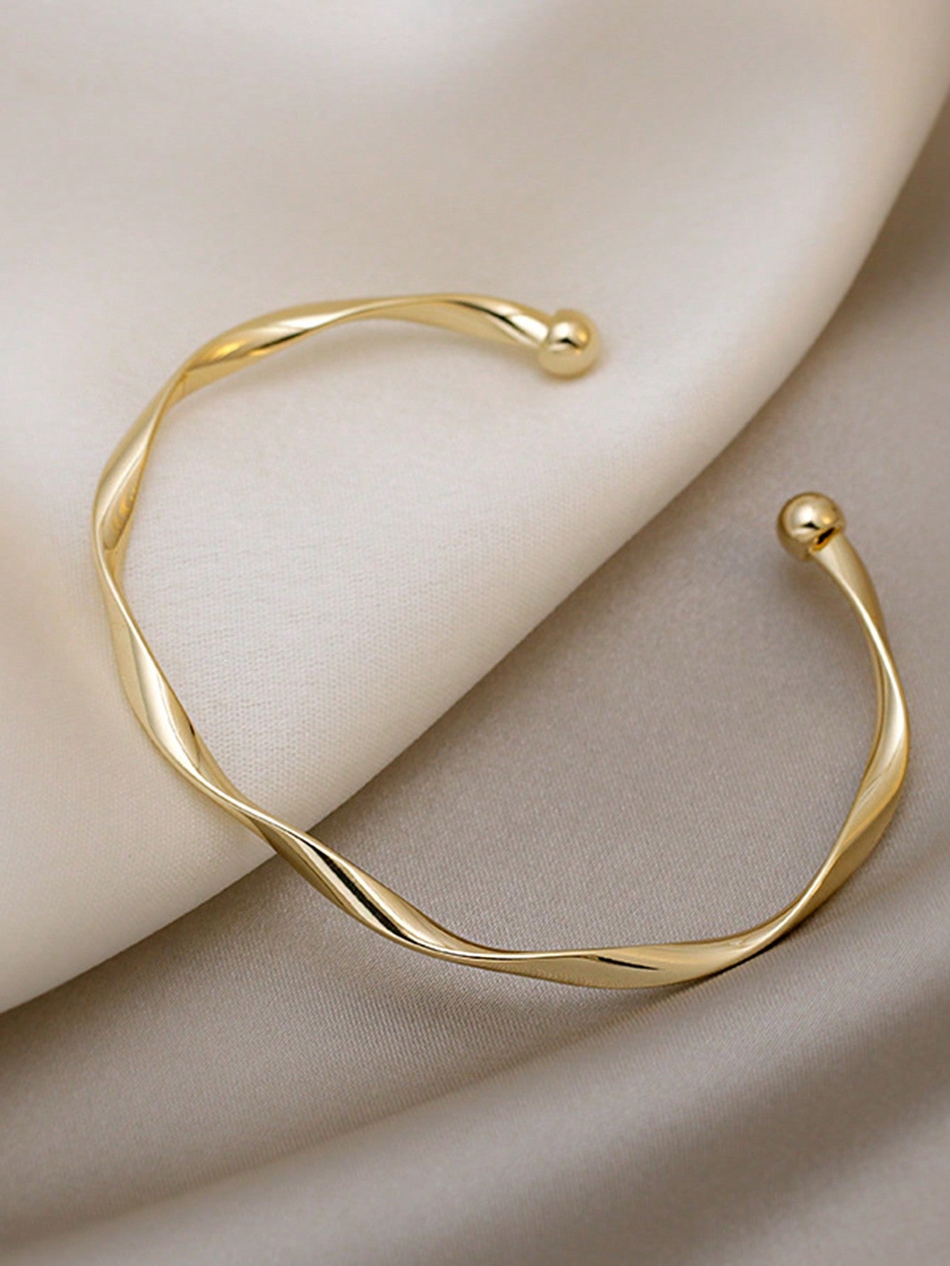 Gold twisted bangle bracelet with a ball accent on one end. Elegant, minimalist design displayed on a gray fabric background