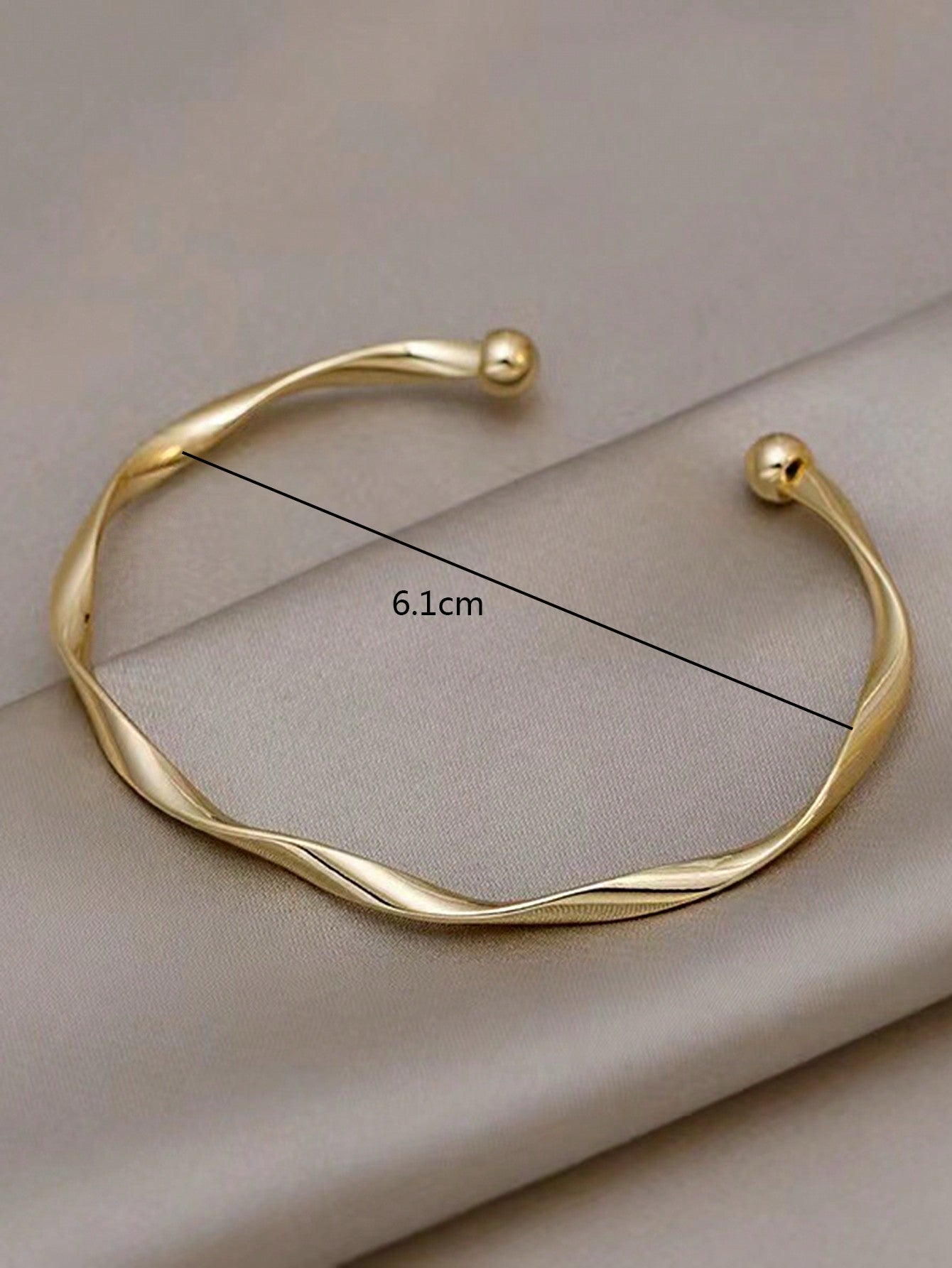 Gold twisted bangle bracelet with a ball accent on one end. Elegant, minimalist design displayed on a gray fabric background