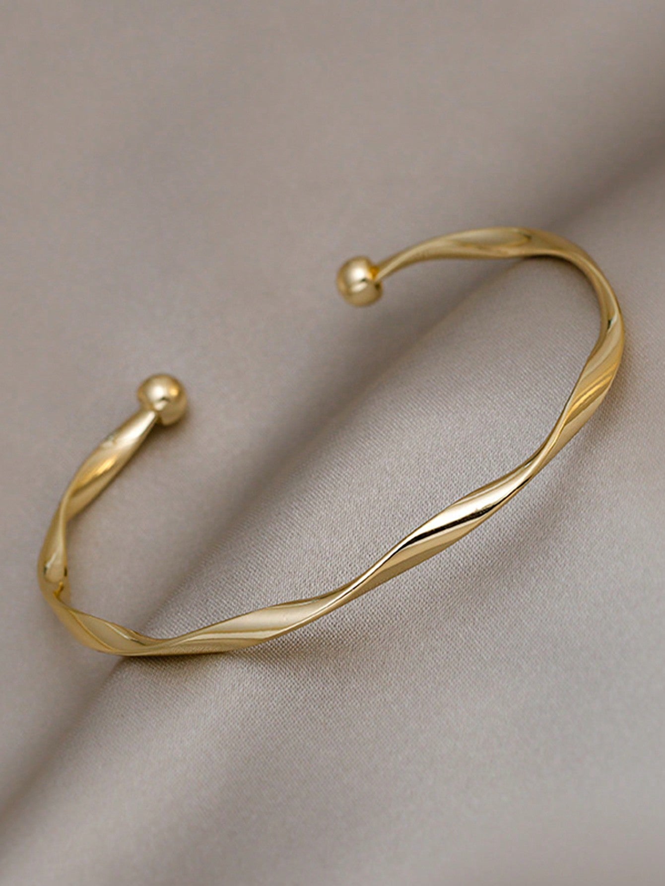 Gold twisted bangle bracelet with a ball accent on one end. Elegant, minimalist design displayed on a gray fabric background