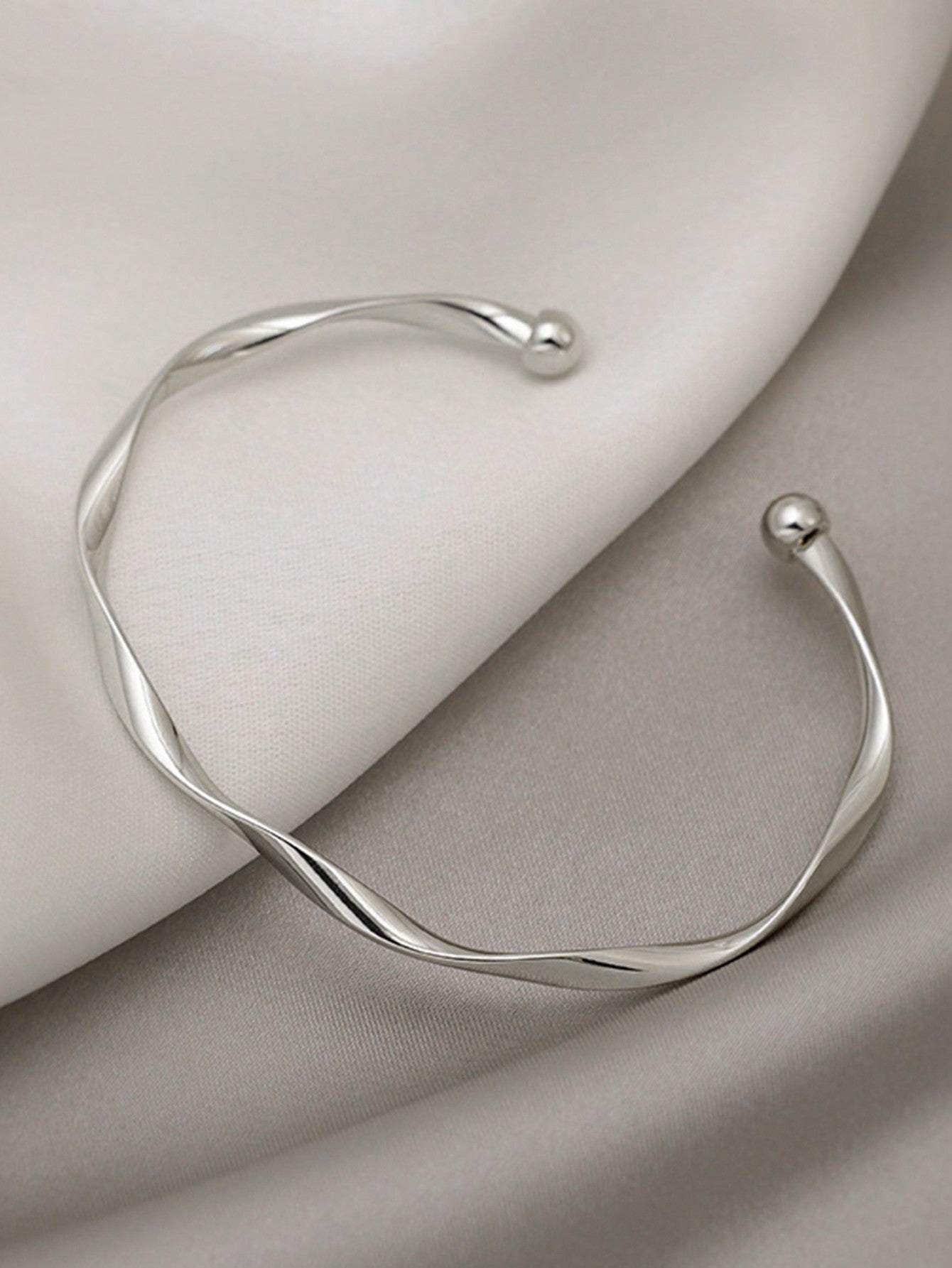 Silver twisted bangle bracelet with a ball accent on one end. Elegant, minimalist design displayed on a gray fabric background