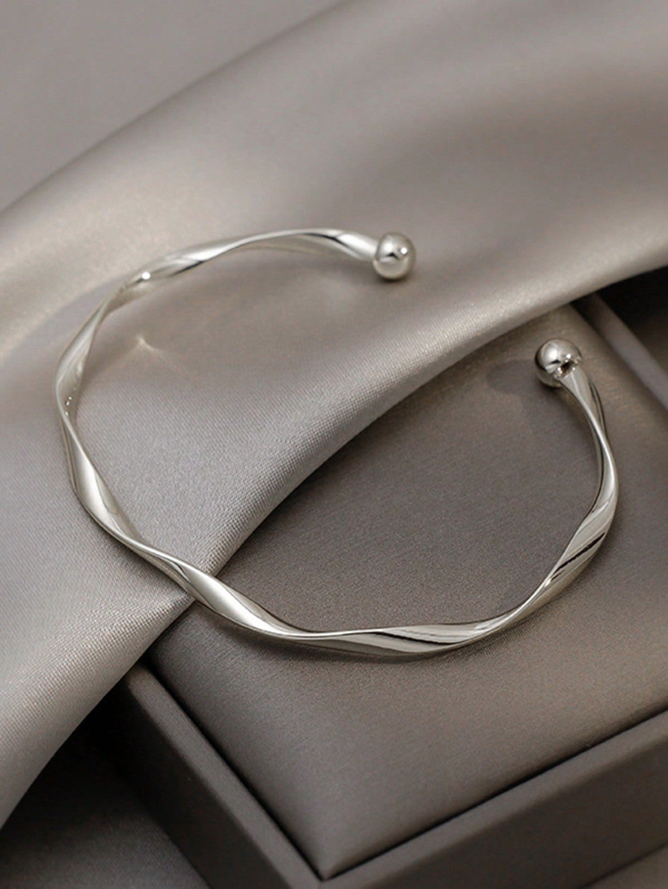 Silver twisted bangle bracelet with a ball accent on one end. Elegant, minimalist design displayed on a gray fabric background