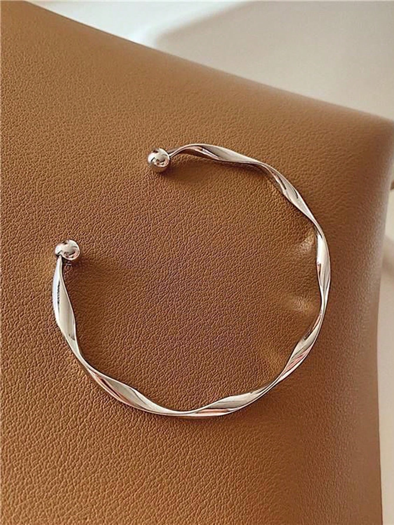 Silver twisted bangle bracelet with a ball accent on one end. Elegant, minimalist design displayed on a gray fabric background