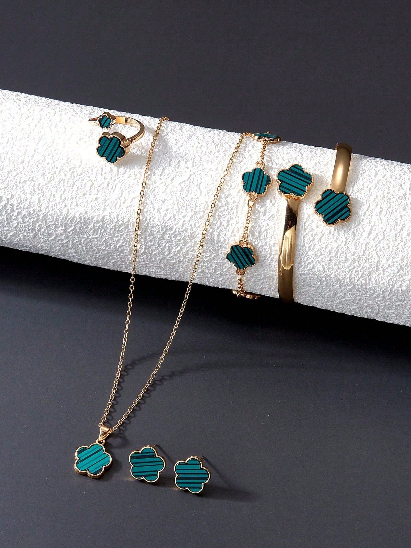 Gold and Green flower-shaped jewelry set including a necklace, bracelet, bangle, earrings, and ring. Arranged on a white textured surface against a dark background.
