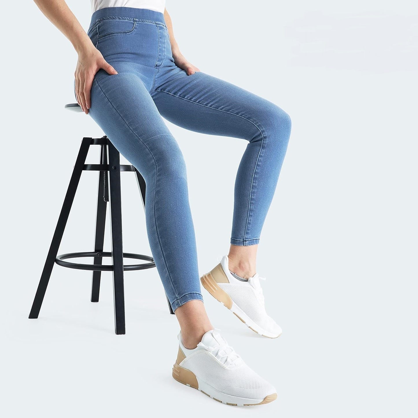 Woman wearing Blue-Wash high-waisted skinny jeans with functional back pockets, paired with a white short-sleeved shirt and white sneakers while sitting on a stool.