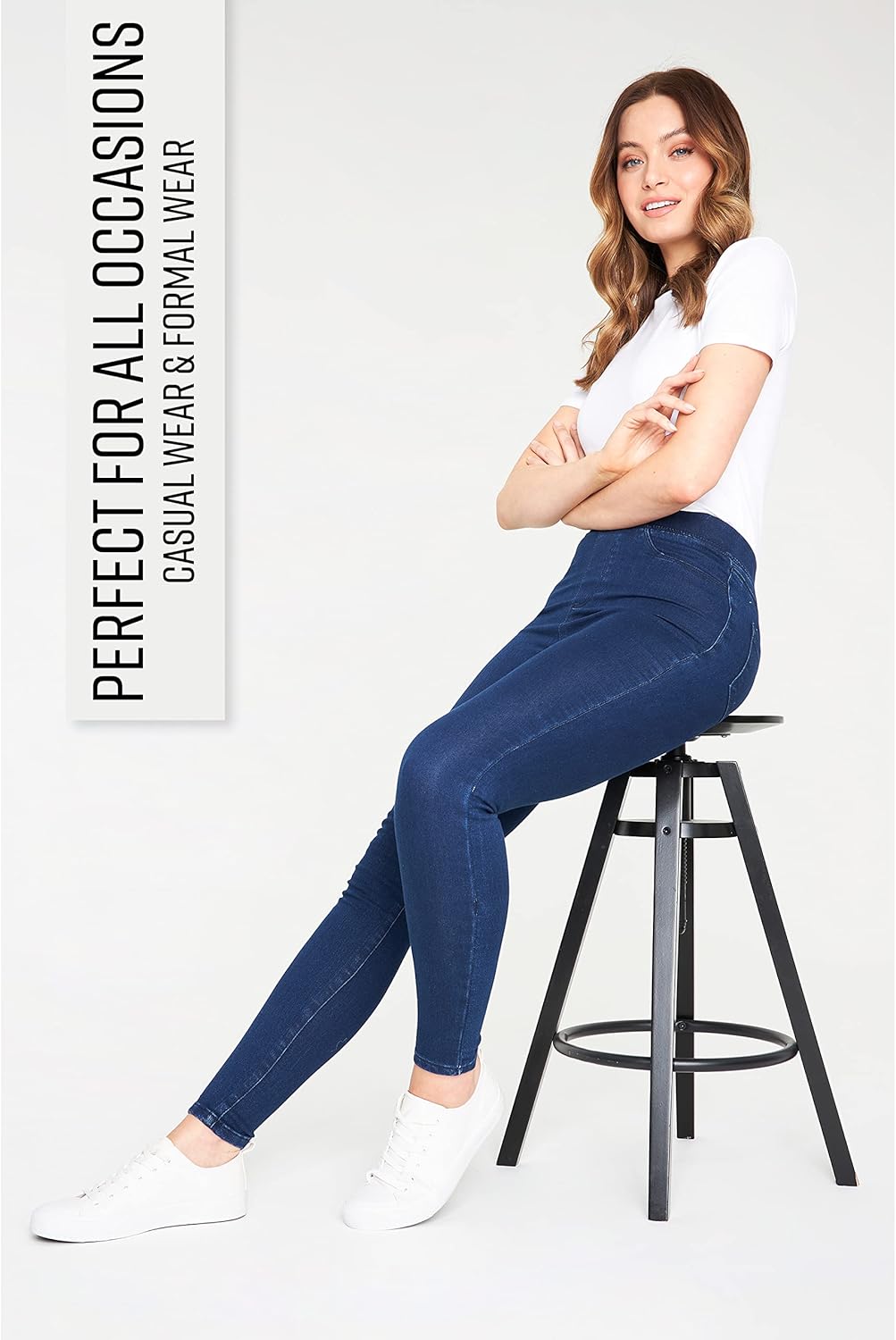 Woman wearing dark blue leggings with pockets, a white short-sleeved shirt, and white sneakers while sitting on a stool.