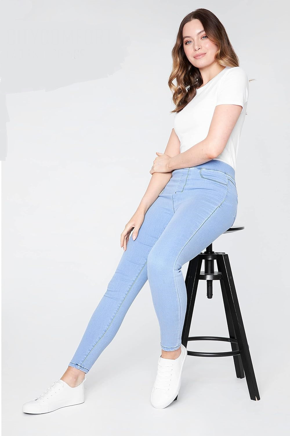 Woman wearing light blue skinny jeans with a white short-sleeved shirt and white sneakers while sitting on a stool.