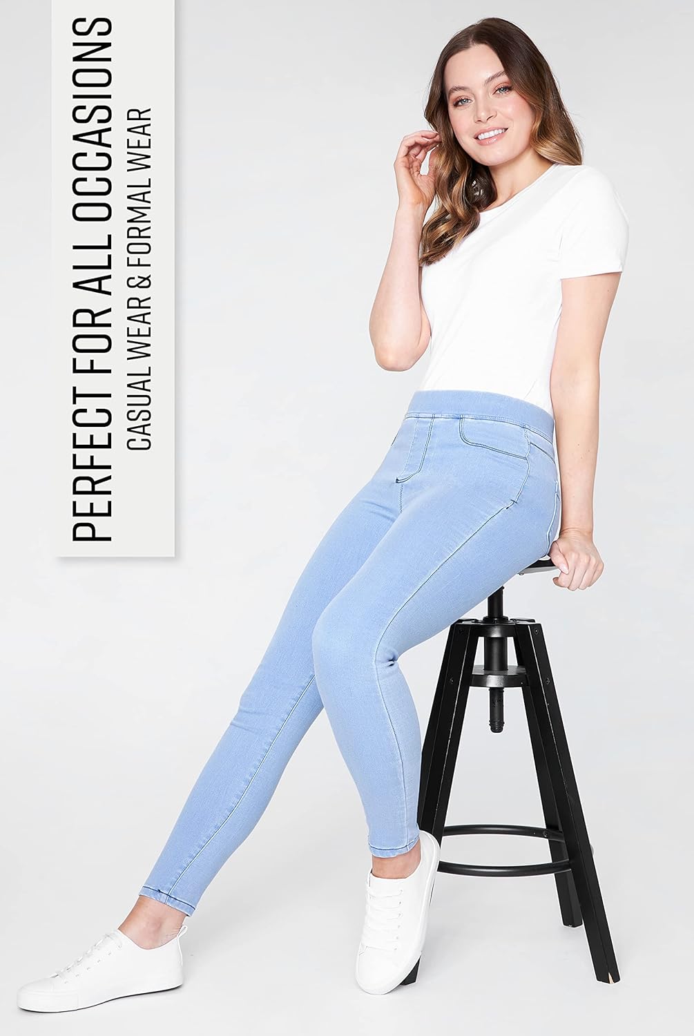 Woman wearing light blue high-waisted skinny jeans with a white short-sleeved shirt and white sneakers while sitting on a stool.
