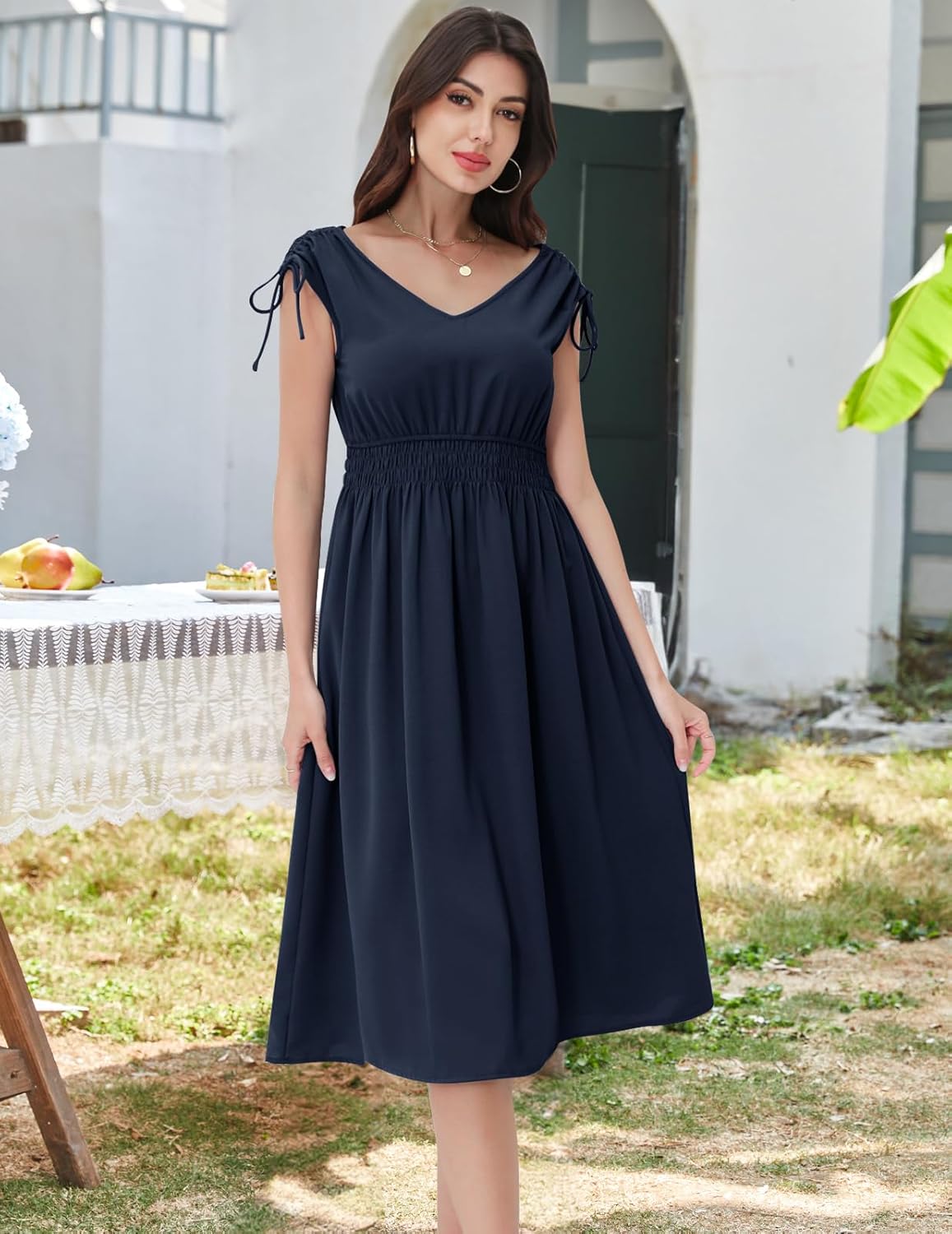 Navy midi dress with a V-neckline, short sleeves with drawstring ties, elasticated waistband, and a flowy skirt. Paired with gold hoop earrings and a delicate necklace. Shown in an outdoor setting with greenery and a table with food items.