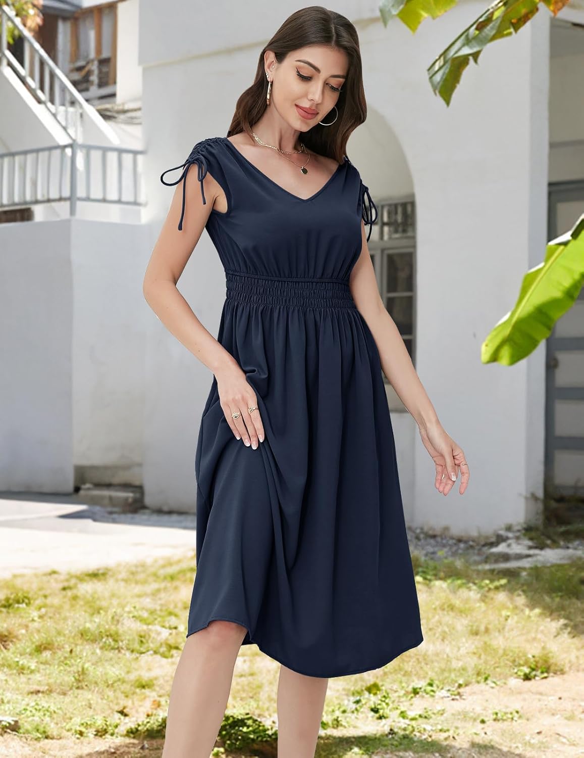 Navy dress with a V-neckline, short sleeves with drawstring ties, elasticated waistband, and a flowy skirt. Paired with gold hoop earrings and a delicate necklace. Shown in an outdoor setting with greenery and architecture.