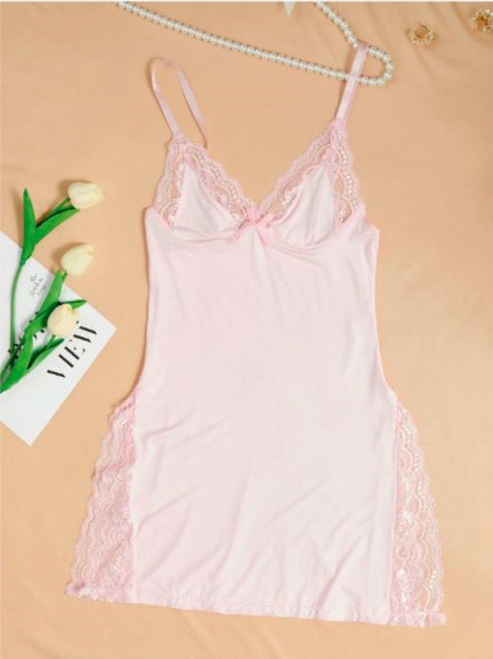 Pink chemise with lace trim and thin straps. The garment is displayed on a hanger against a peach background, accompanied by a card with "V-Day" written on it and a few white tulips.