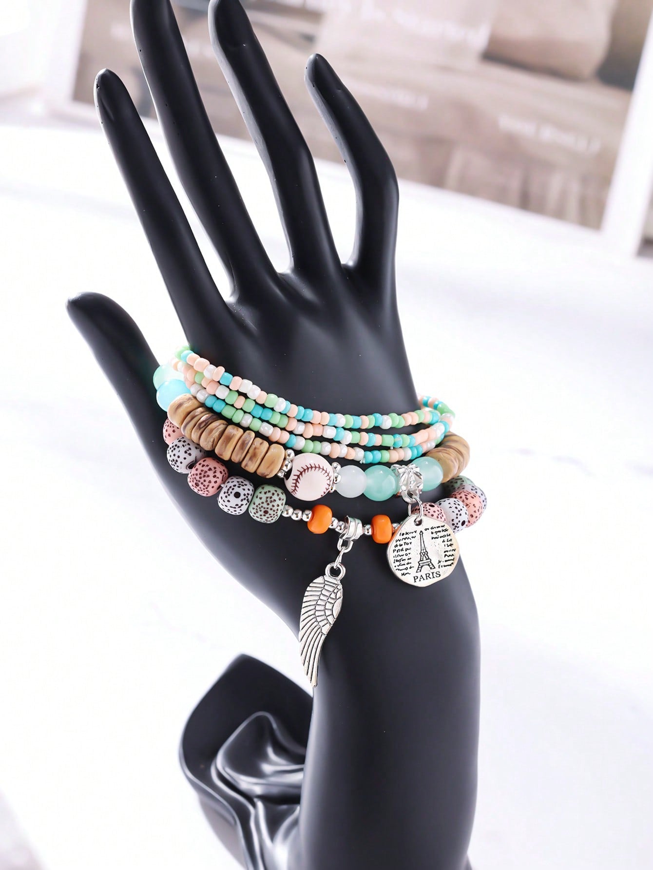 Stacked pastel-colored bracelet set featuring mint green, peach, and white beads with wood accents. Includes clear bands and patterned beads. Charm details include a metallic wing pendant and round Eiffel Tower medallion engraved with 'PARIS' and script text. Versatile design for layering or gifting.