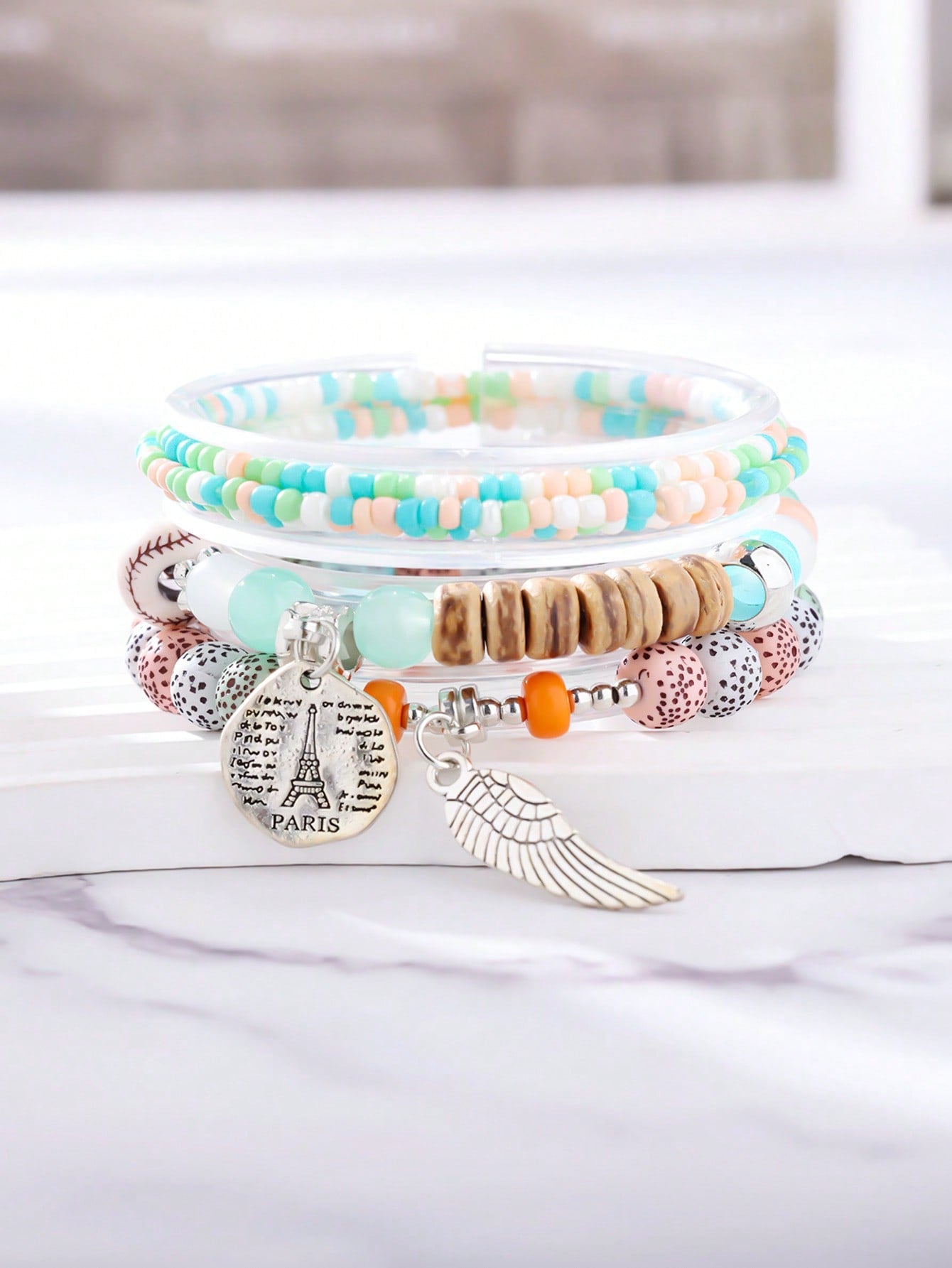 Stacked pastel-colored bracelet set featuring mint green, peach, and white beads with wood accents. Includes clear bands and patterned beads. Charm details include a metallic wing pendant and round Eiffel Tower medallion engraved with 'PARIS' and script text. Versatile design for layering or gifting.