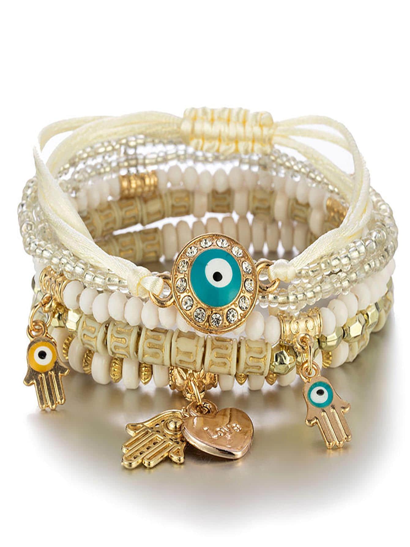Stack of multilayered white beaded bracelets with gold accents, featuring a large turquoise evil eye pendant, gold Hamsa hand charms, and small bead details. Bohemian-inspired design with adjustable ties for a personalized fit