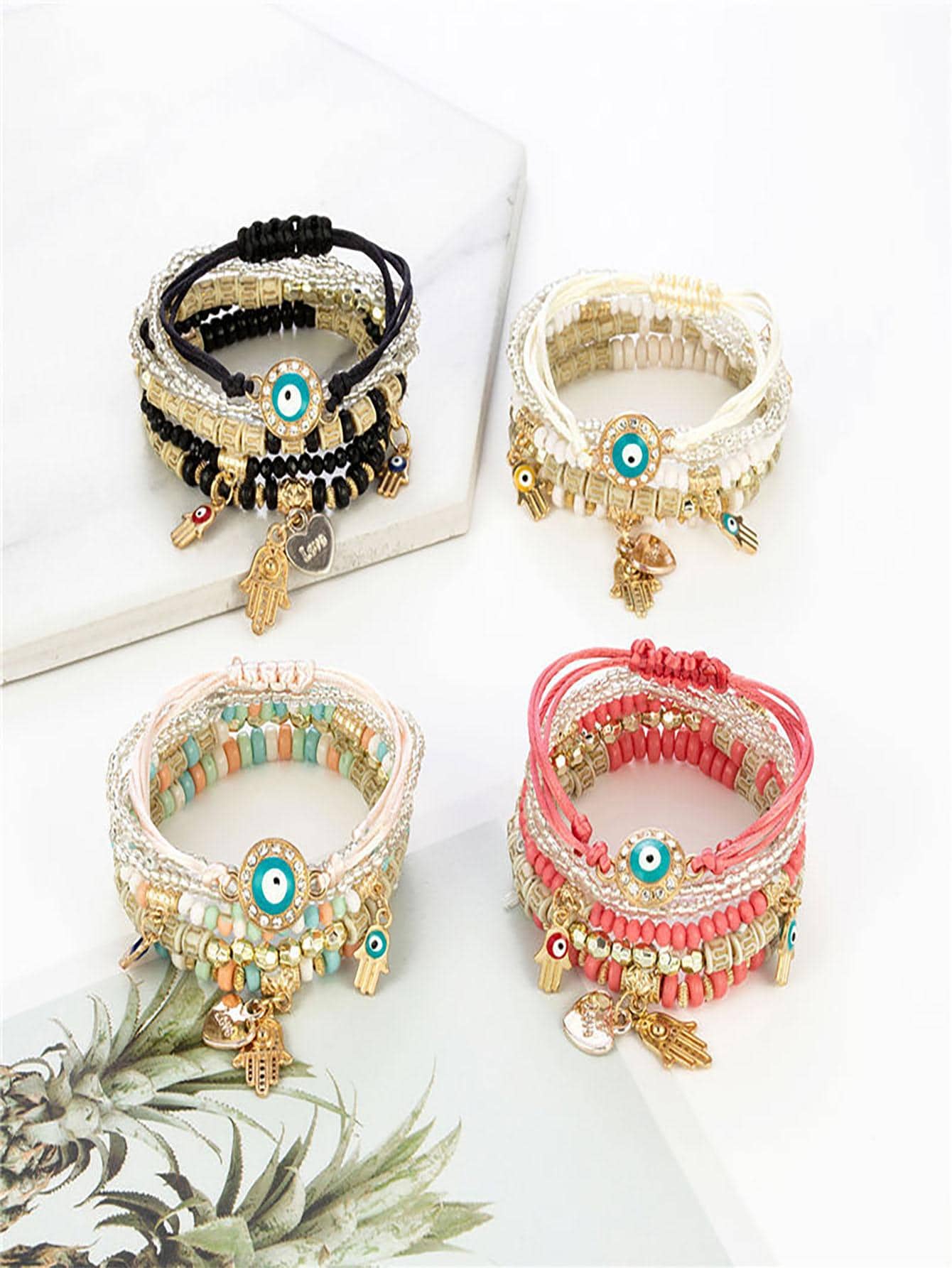 Set of four multilayered beaded bracelets in black, white, pink, and multicolor. Each features small beads, gold accents, and charm details like evil eye pendants and Hamsa hands. Bohemian-inspired design with adjustable ties.