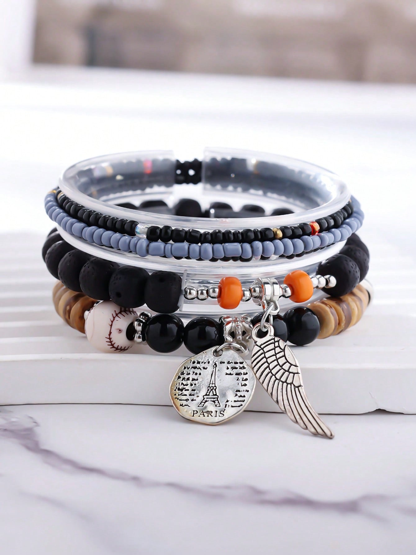 Stack of multicolored beaded bracelets featuring blue, white, and wooden beads with gold accents. Includes a silver wing charm and a round pendant engraved with 'PARIS'. Layered design for a vibrant, travel-inspired look.