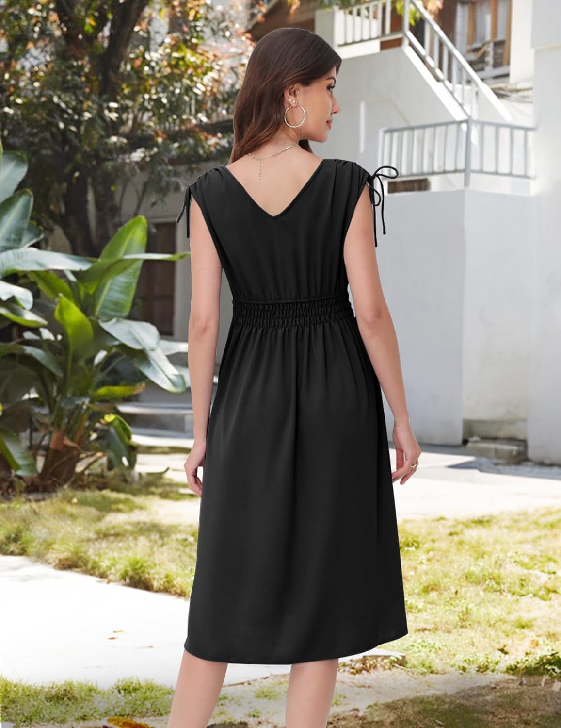 Black midi dress with a V-neckline at the back, short sleeves with drawstring ties, elasticated waistband, and a flowy skirt. Shown from the back in an outdoor setting with greenery and architecture.