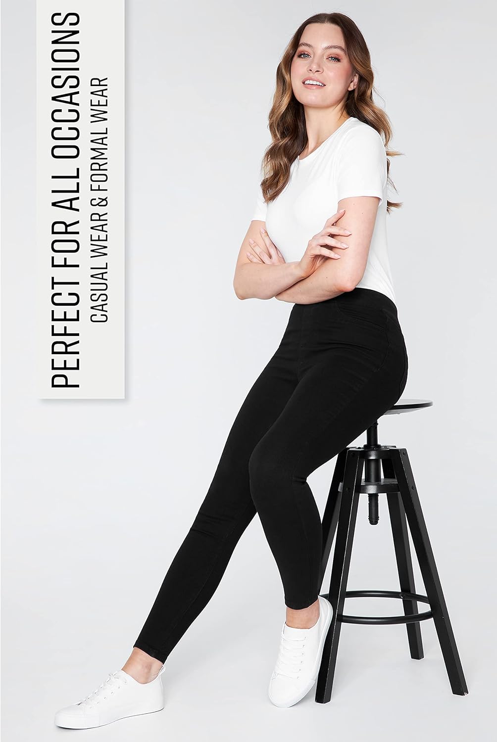 Woman wearing black high-waisted stretchy leggings with functional back pockets, paired with a white short-sleeved shirt and white sneakers while sitting on a stool.