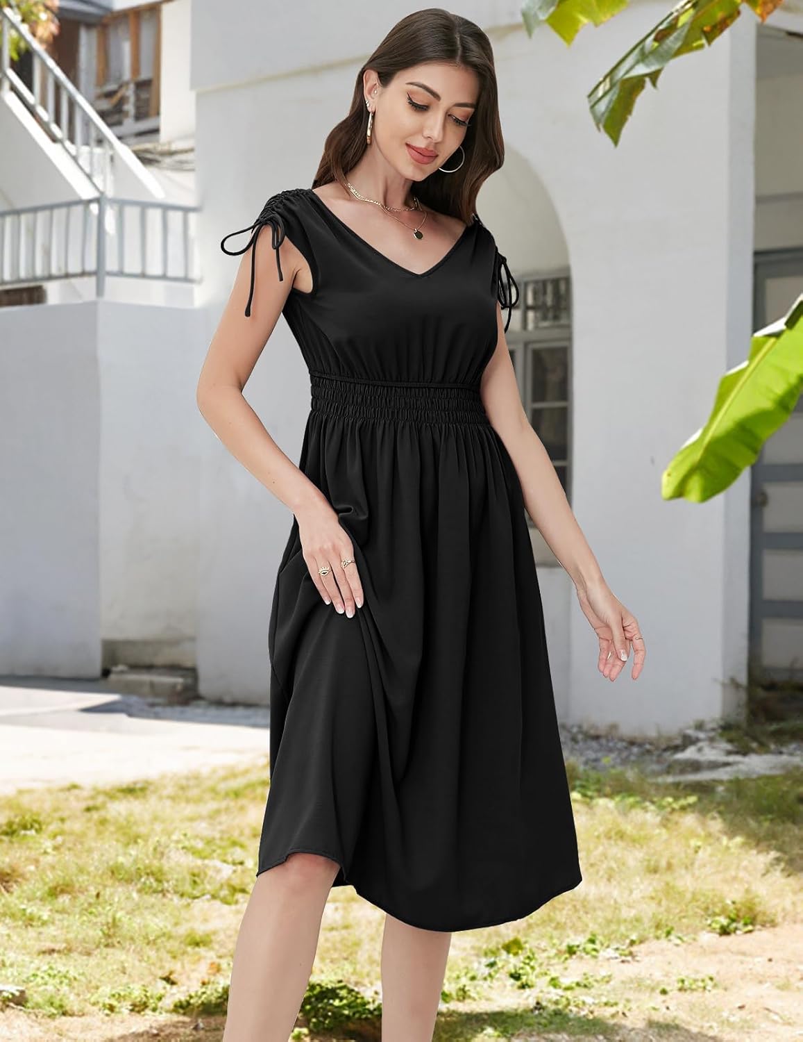 Black V-neck midi dress with elasticated waistband, short ruched sleeves with drawstring ties, and a flowy skirt, paired with gold hoop earrings and a delicate necklace, shown in an outdoor setting with greenery and architecture.
