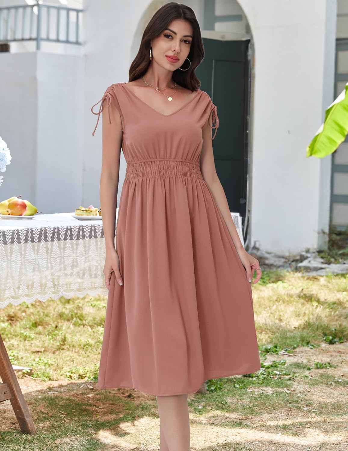 Blush  dress with a V-neckline, short sleeves with drawstring ties, elasticated waistband, and a flowy skirt. Paired with gold hoop earrings and a delicate necklace. Shown in an outdoor setting with greenery and a table with food items.