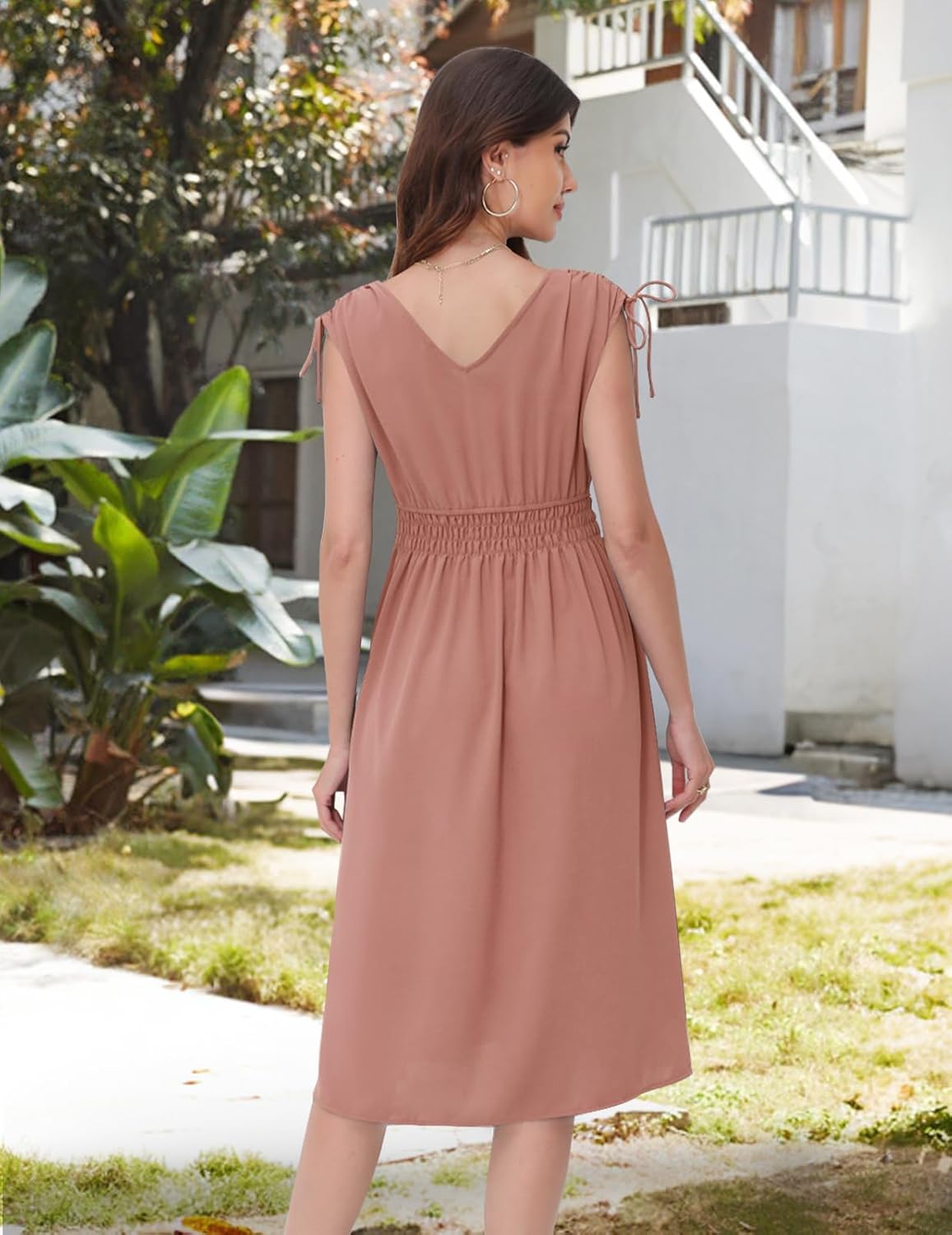 Blush dress with a V-neckline at the back, short sleeves with drawstring ties, elasticated waistband, and a flowy skirt. Paired with gold hoop earrings and a delicate necklace. Shown from the back in an outdoor setting with greenery and architecture.