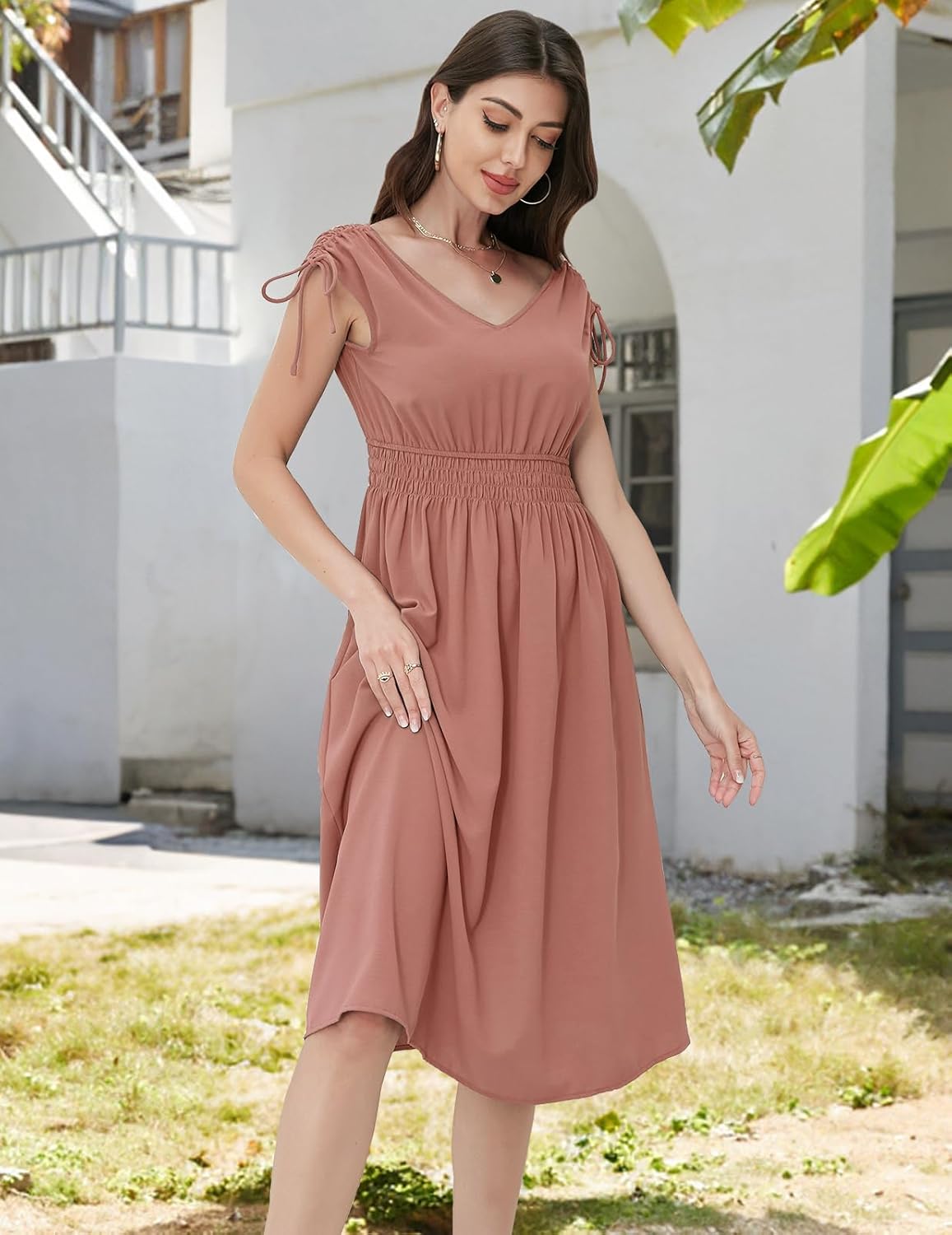 Blush dress with a V-neckline, short sleeves with drawstring ties, elasticated waistband, and a flowy skirt. Paired with gold hoop earrings and a delicate necklace. Shown in an outdoor setting with greenery and architecture.