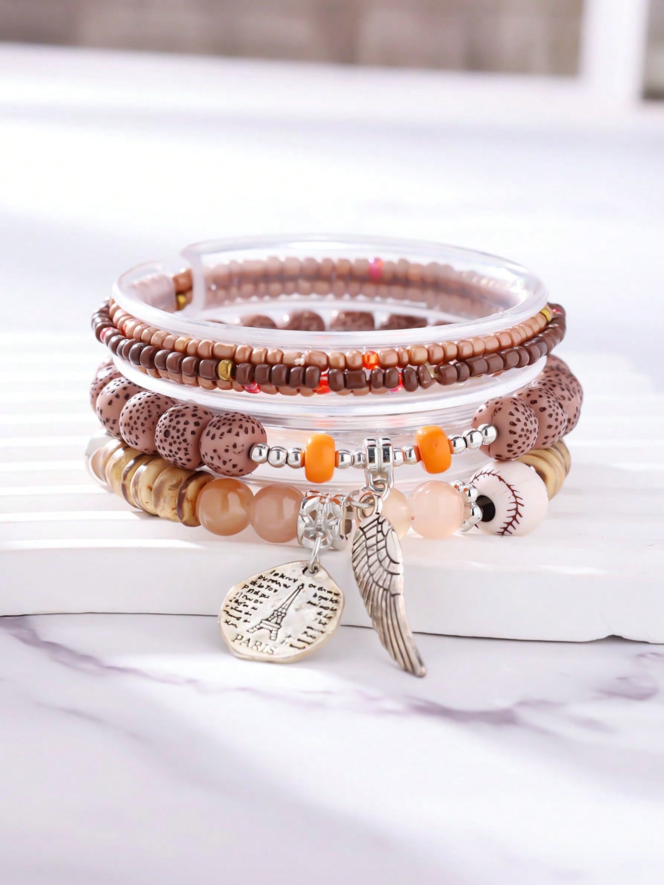 Stacked neutral-toned bracelet set with brown, beige, and transparent beads. Includes wood accents, patterned beads, and clear bands. Features a metallic wing pendant and round Eiffel Tower charm engraved with 'PARIS' and script text. Versatile design for layering or gifting.