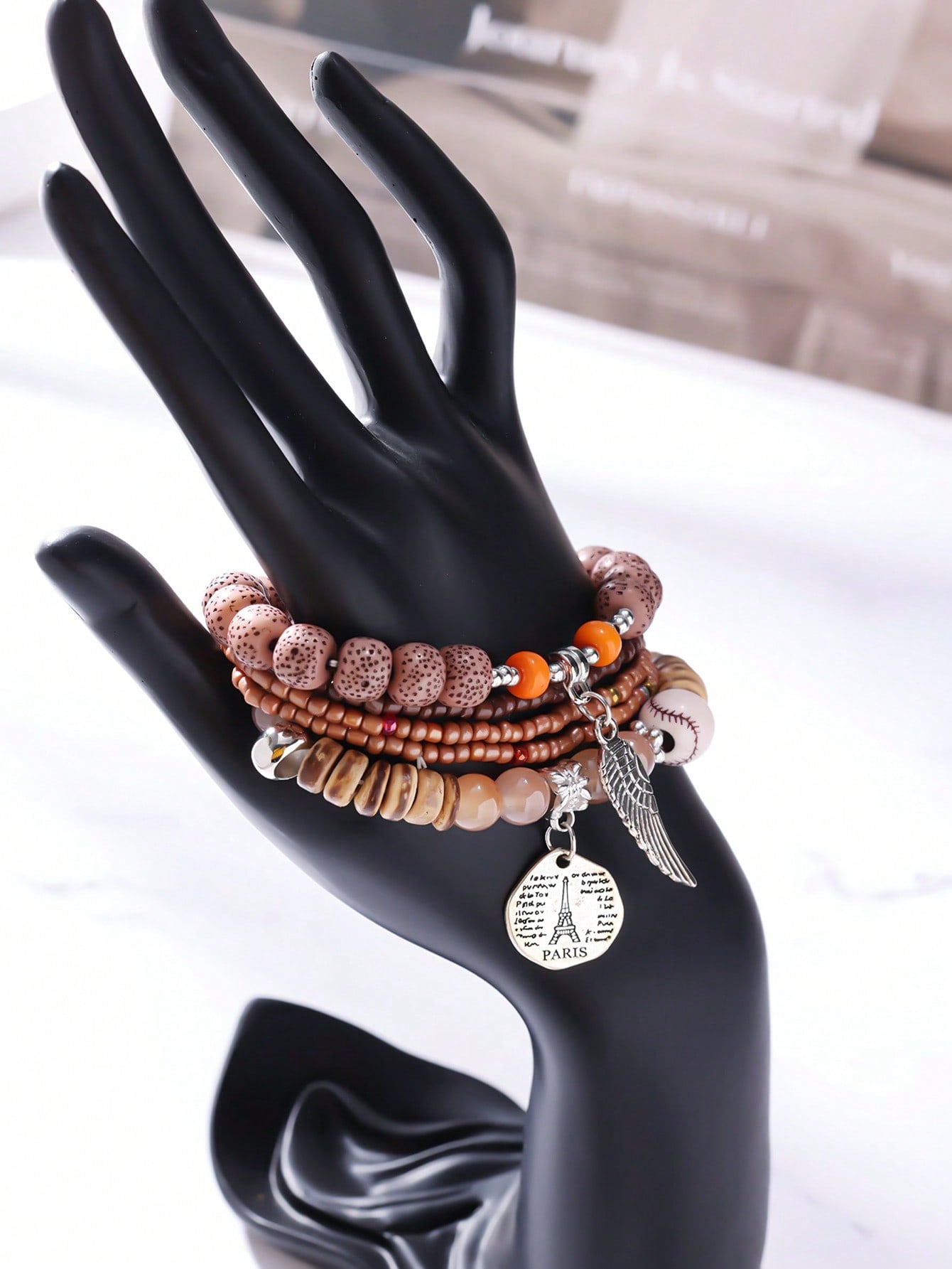 Stacked neutral-toned bracelet set with brown, beige, and transparent beads. Includes wood accents, patterned beads, and clear bands. Features a metallic wing pendant and round Eiffel Tower charm engraved with 'PARIS' and script text. Versatile design for layering or gifting.