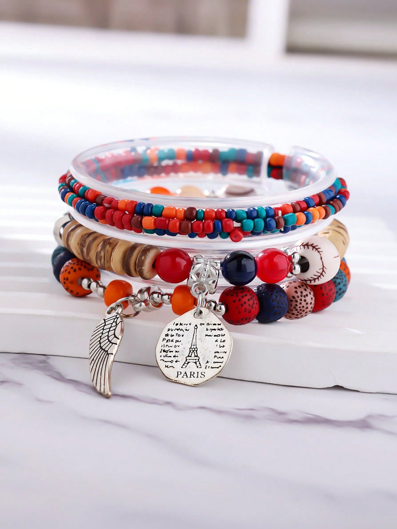 Stacked multicolor bracelet set with red, blue, and transparent beads. Includes wood accents, patterned beads, and clear bands. Features a metallic wing pendant and round Eiffel Tower charm engraved with 'PARIS' and script text. Versatile design for layering or gifting.