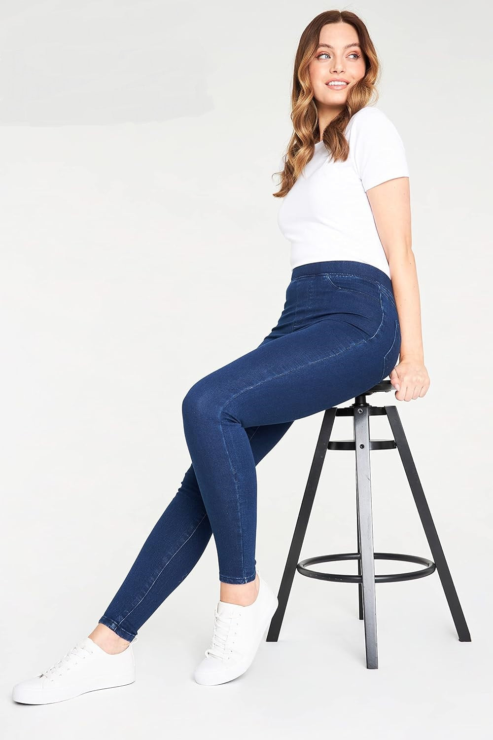 Woman wearing white short-sleeved t-shirt and dark blue skinny jeans with white sneakers, sitting on a black stool against a white background.