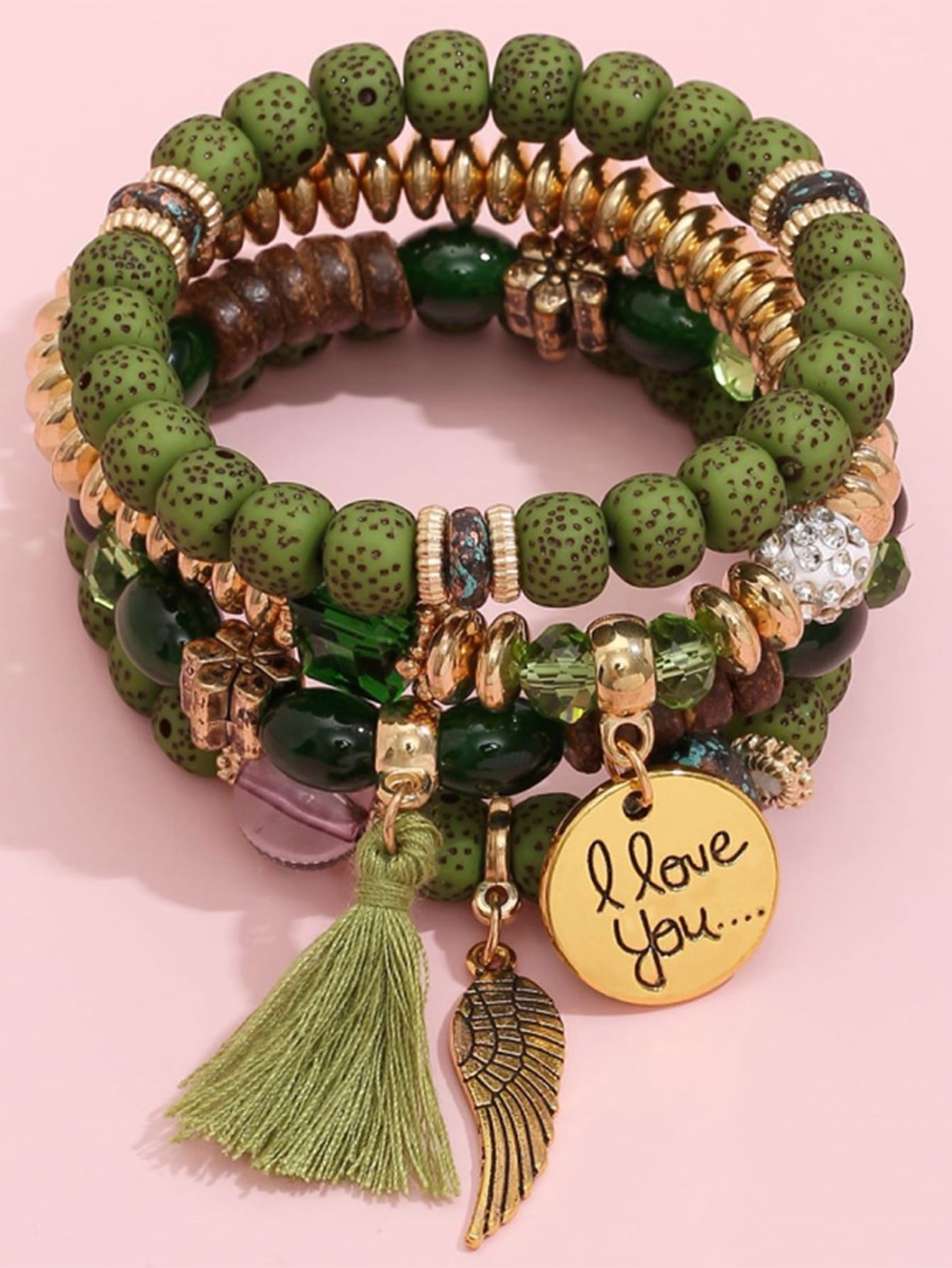 Stack of green beaded bracelets with gold accents featuring a wing charm, gold pendant with 'I love you' text, and a green tassel. Layered design with varied bead textures for an earthy, bohemian style.