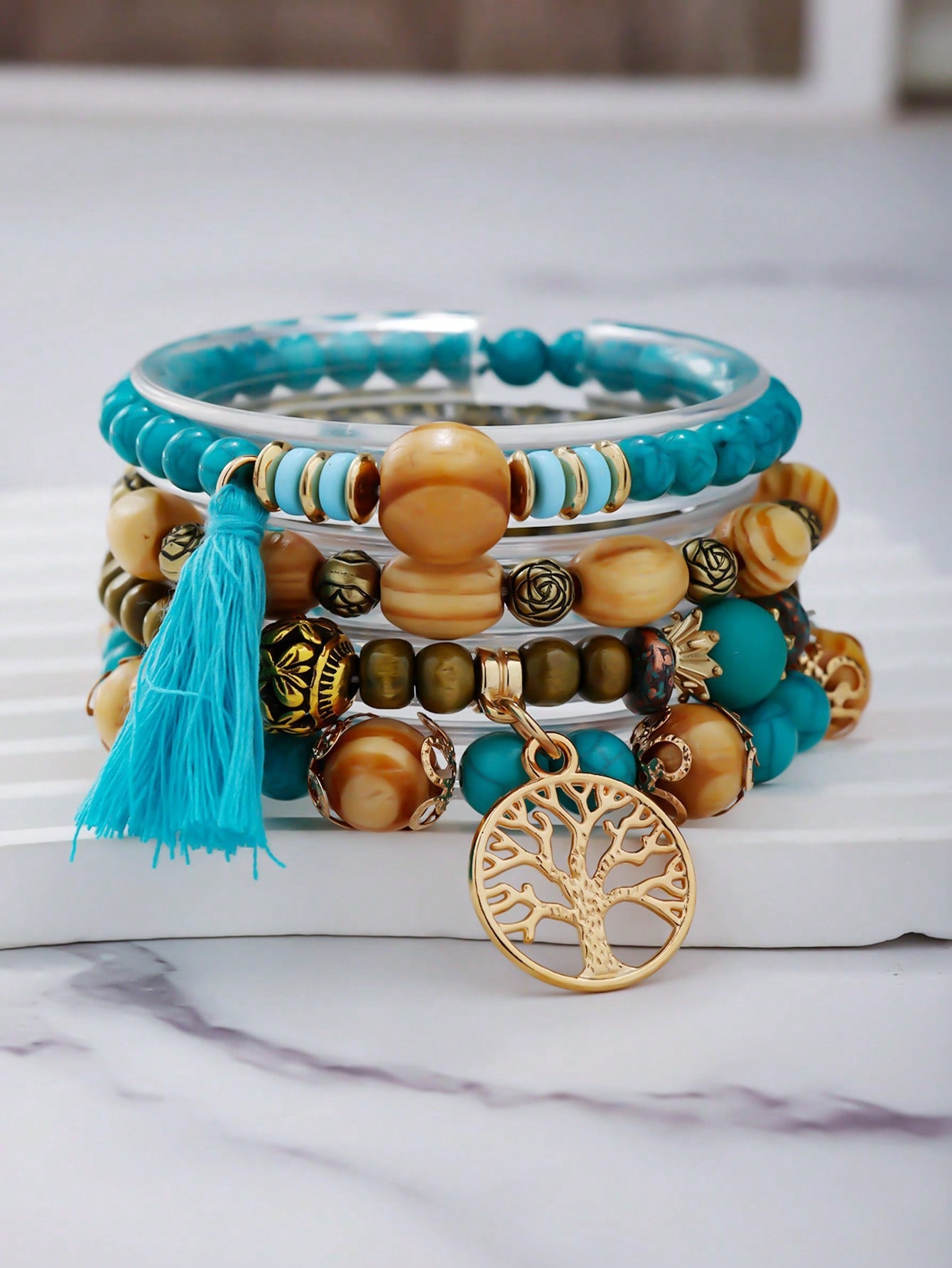 Stacked turquoise and gold bracelet set with clear bands, wooden beads, and decorative metal beads. Includes a turquoise tassel pendant and a large gold tree-of-life medallion charm. Versatile design for layering or gifting.