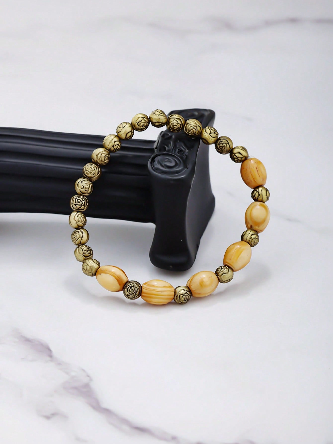 Looped bracelet with alternating gold and light brown beads placed on a black carved holder. Features metallic and wood-like bead finishes.