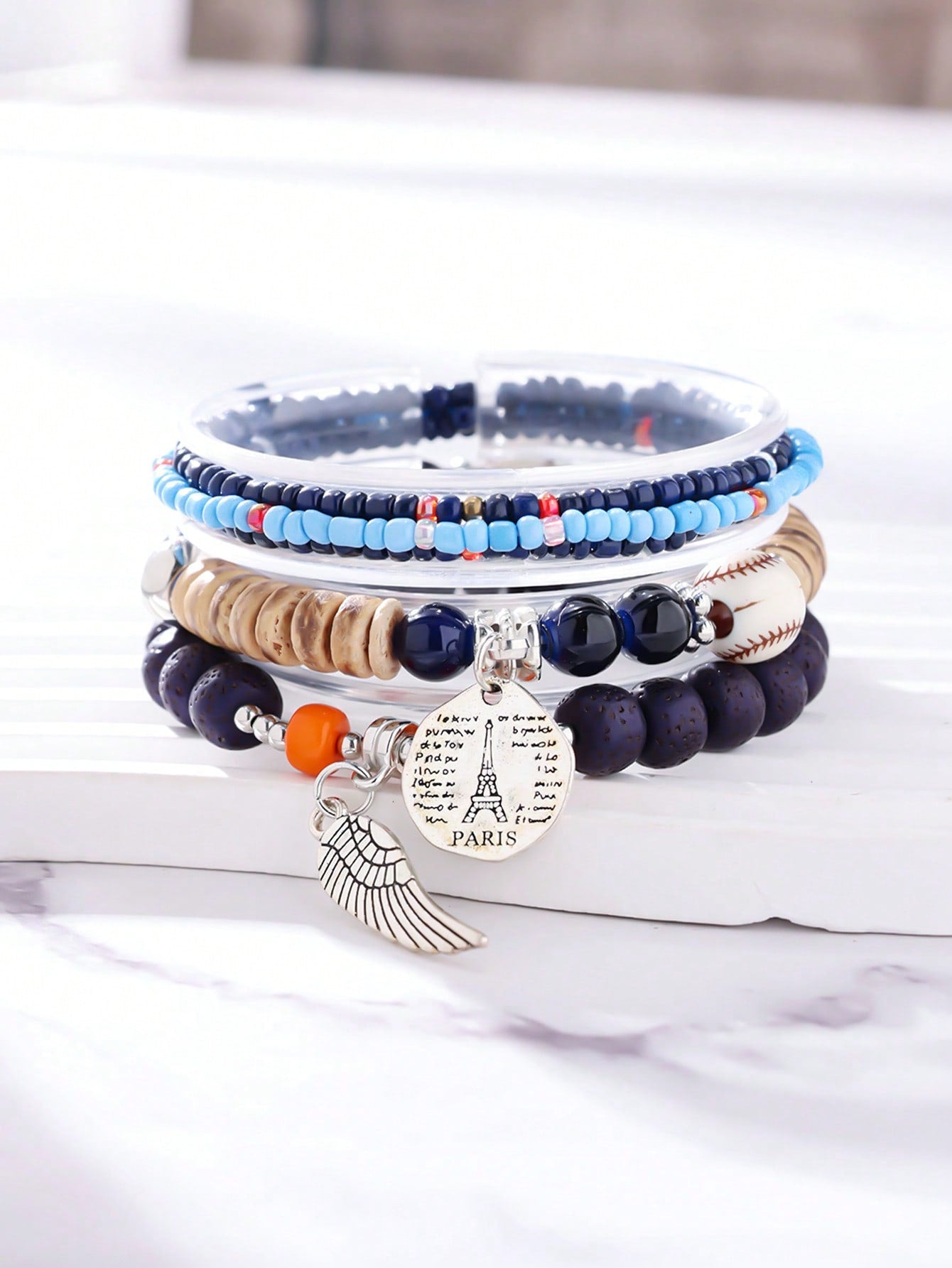 Stacked navy blue and light blue bracelet set with wood beads, transparent bands, and baseball-stitch accent. Features a metallic wing pendant and round Eiffel Tower charm engraved with 'PARIS' and script text. Versatile design for layering with adjustable charms. Ideal for travel-inspired jewelry collections or gift sets.