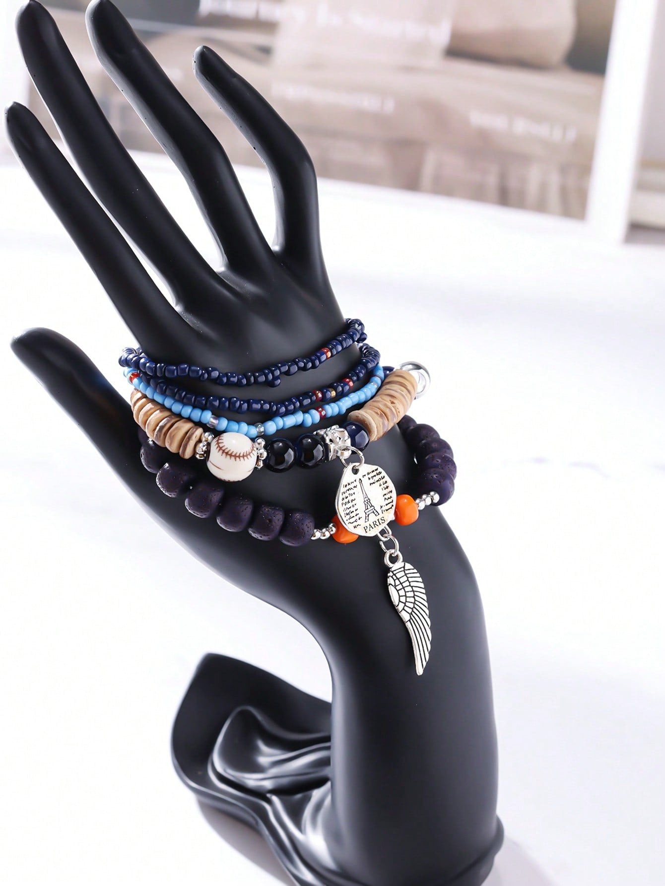 Stacked navy blue and light blue bracelet set with wood beads, transparent bands, and baseball-stitch accent. Features a metallic wing pendant and round Eiffel Tower charm engraved with 'PARIS' and script text. Versatile design for layering with adjustable charms. Ideal for travel-inspired jewelry collections or gift sets.