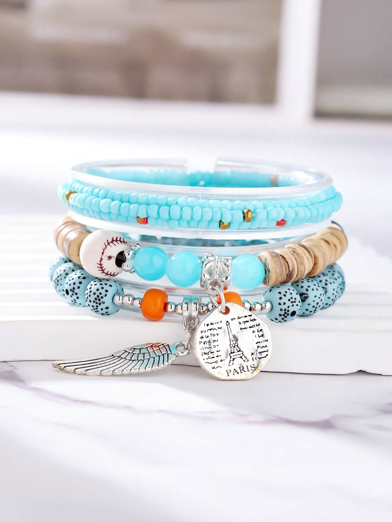 Stackable turquoise and white beaded bracelet set with gold accents, wood beads, Eiffel Tower charm, and angel wing pendant. Versatile layered design for travel-inspired style. Features mixed textures and charms perfect for stacking or gifting.