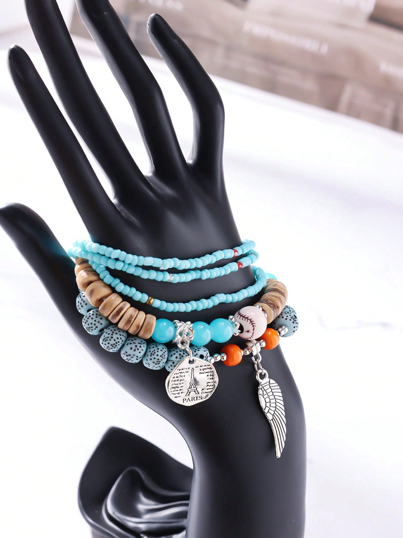 Stackable turquoise and white beaded bracelet set with gold accents, wood beads, Eiffel Tower charm, and angel wing pendant. Versatile layered design for travel-inspired style. Features mixed textures and charms perfect for stacking or gifting.