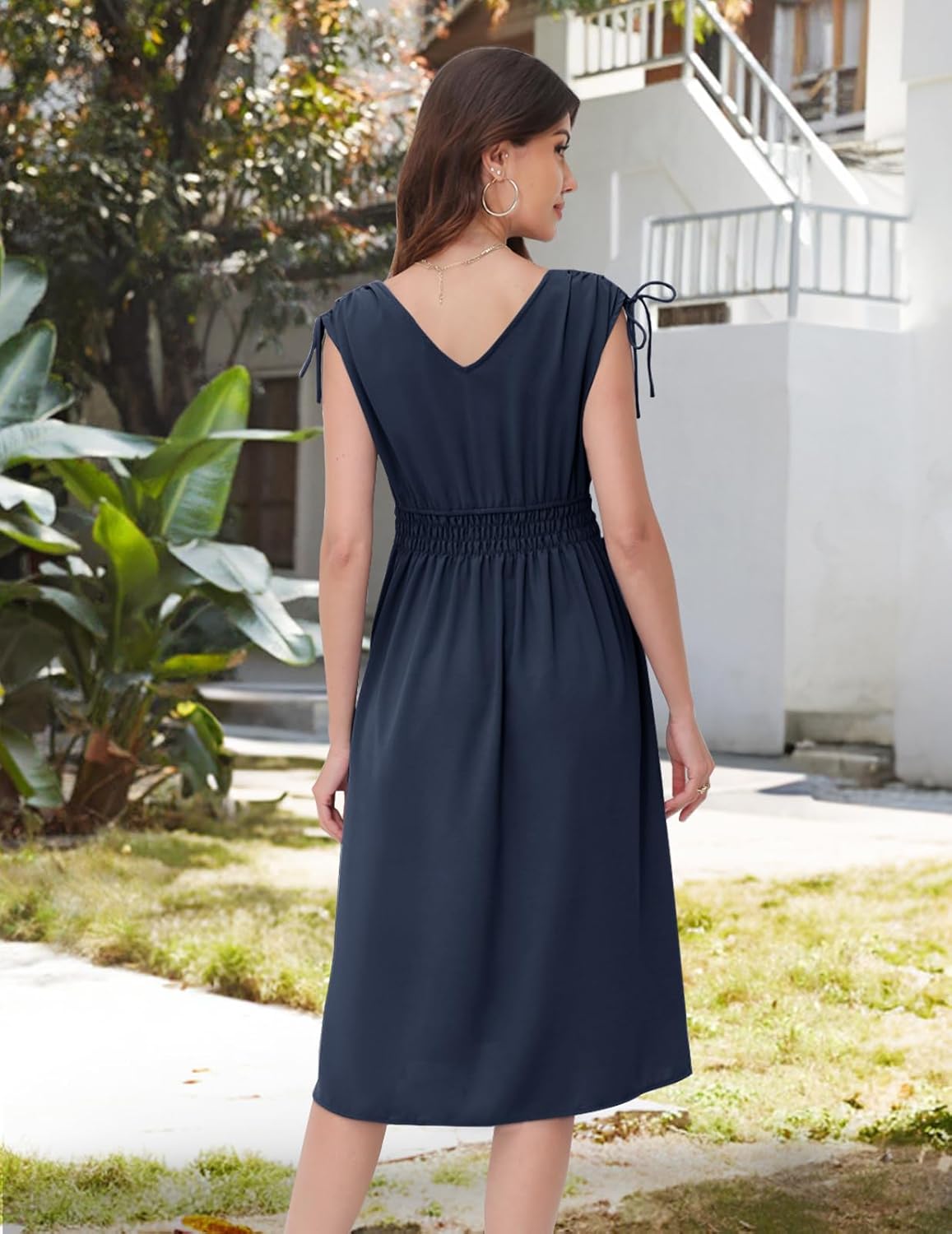 Navy dress with a V-neckline at the back, short sleeves with drawstring ties, elasticated waistband, and a flowy skirt. Paired with gold hoop earrings and a delicate necklace. Shown from the back in an outdoor setting with greenery and architecture.
