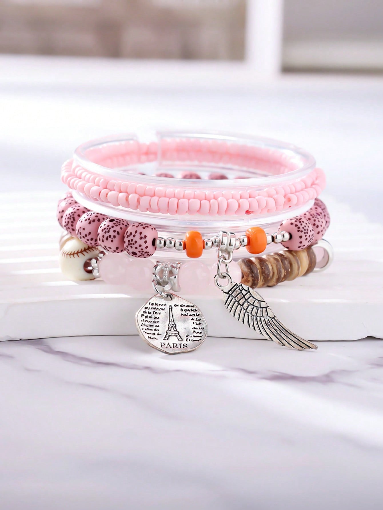 Stacked pink and transparent bracelet set with solid pink and patterned beads. Includes wood accents, clear bands, and a baseball-stitch accent bead. Features a metallic wing pendant and round Eiffel Tower charm engraved with 'PARIS' and script text. Versatile design for layering or gifting.