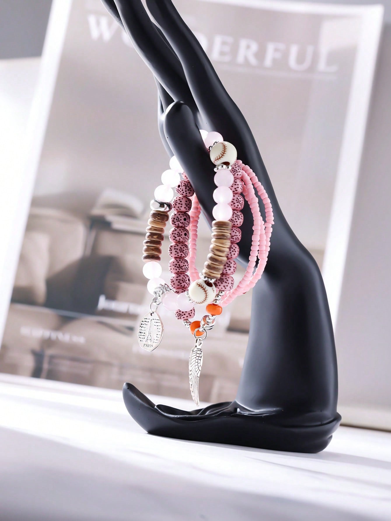 Stacked pink and transparent bracelet set with solid pink and patterned beads. Includes wood accents, clear bands, and a baseball-stitch accent bead. Features a metallic wing pendant and round Eiffel Tower charm engraved with 'PARIS' and script text. Versatile design for layering or gifting.