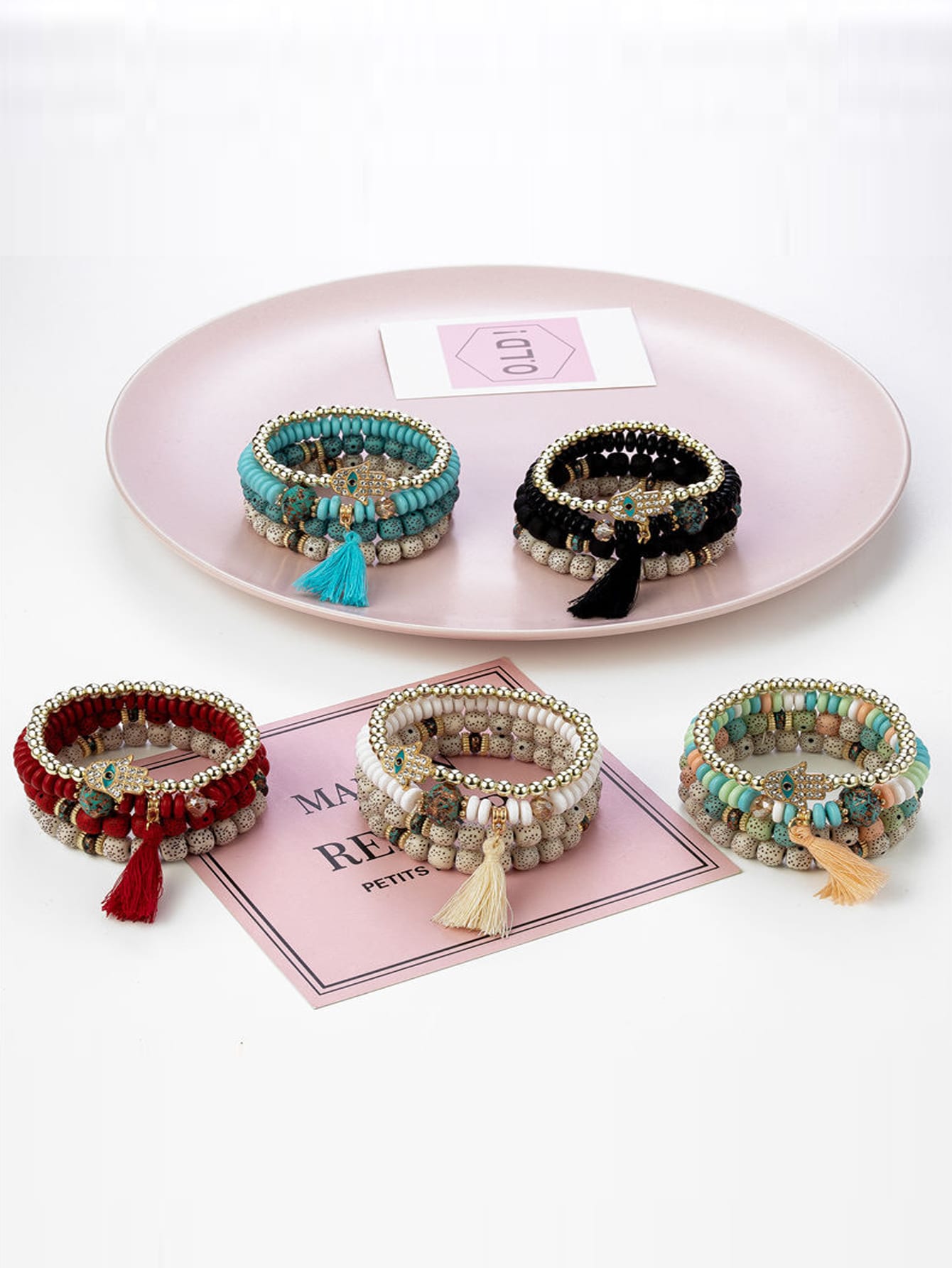 Collection of six beaded bracelets in assorted colors including red, blue, black, white, and multi-color with gold accents. Each features unique designs with beads, tassels, and charm details. Displayed on a pink tray with decorative cards.
