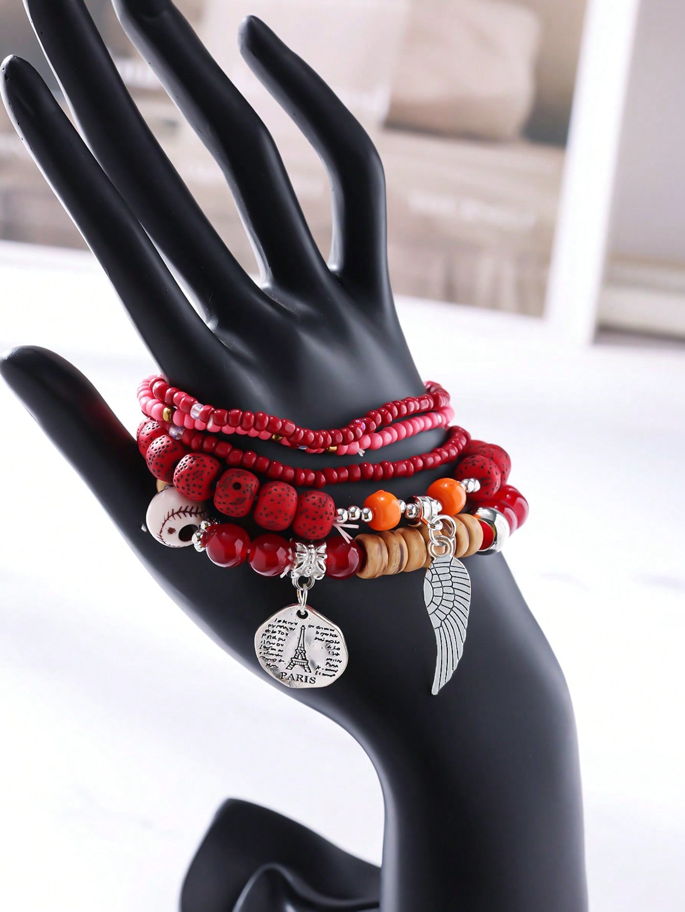 Stack of multicolored beaded bracelets in red, pink, black, and clear tones with gold accents. Features a silver wing charm and a round pendant engraved with 'PARIS'. Layered design displayed on a white surface with a pink background.