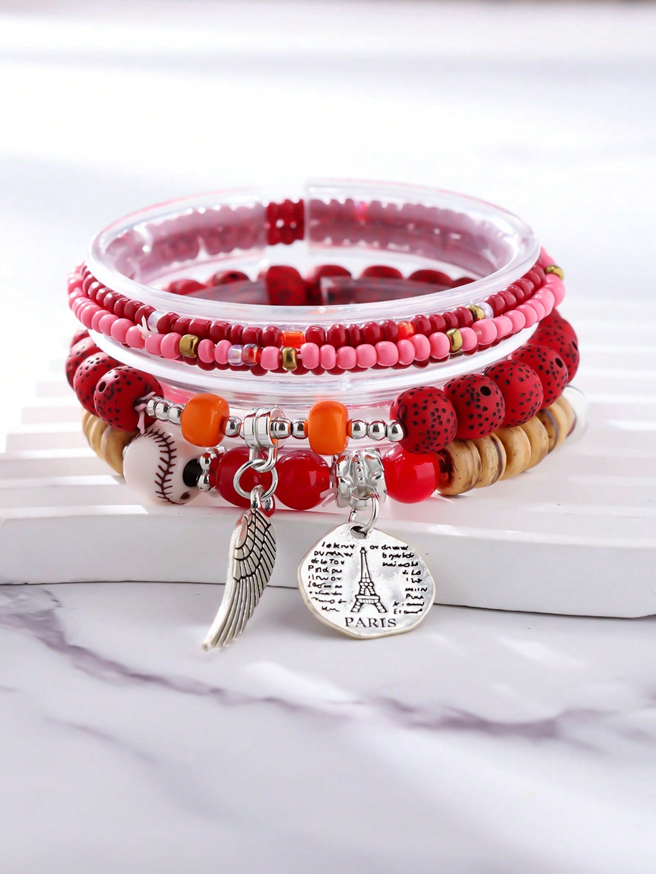 Stack of multicolored beaded bracelets in red, pink, black, and clear tones with gold accents. Features a silver wing charm and a round pendant engraved with 'PARIS'. Layered design displayed on a white surface with a pink background.