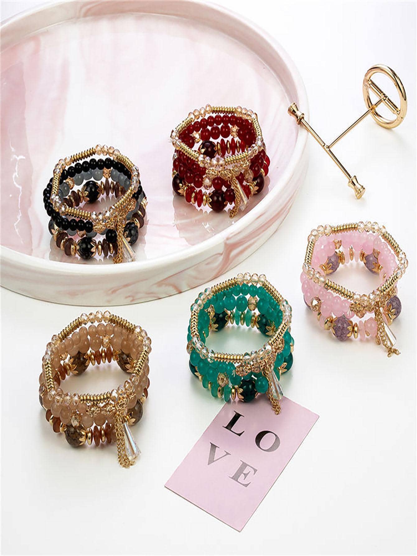 Collection of colorful beaded bracelets in pink, green, black, red, and gold tones. Each features multilayered designs with varying bead sizes, gold accents, and charm details. Displayed on a pink tray with a decorative 'LOVE' card.